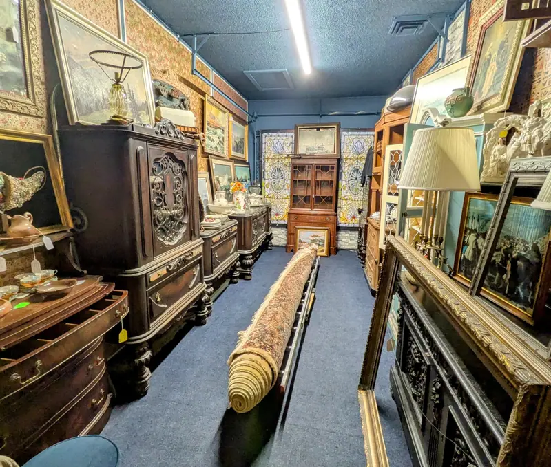 Carved antique sideboards, lamps and framed paintings along a carpeted aisle at Sample Studios