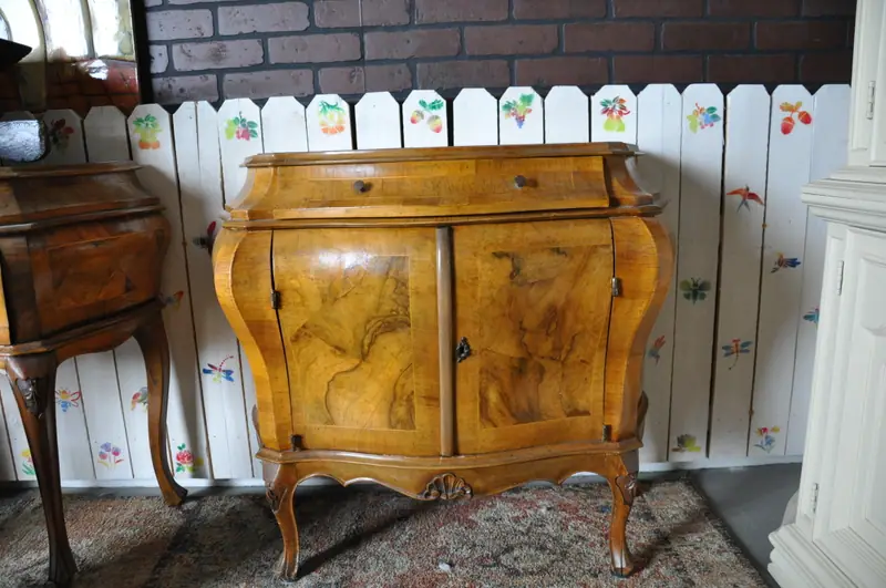 A burl walnut bombe cabinet in front of a painted picket fence at Sample Studios